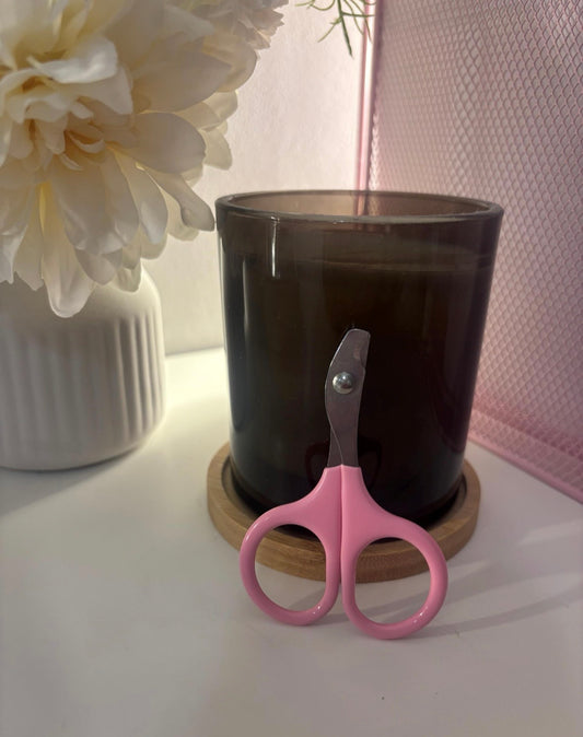 Pink-handled small scissors leaning against a dark amber glass candle on a wooden coaster, styled on a white surface with a white flower vase and pink mesh backdrop.