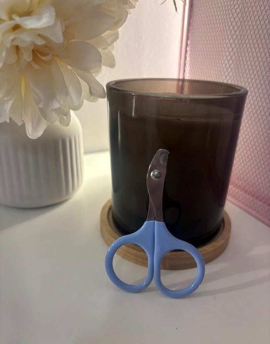 A pair of small nail scissors with light blue handles and silver metal blades, resting in front of a dark amber glass candle on a wooden coaster.